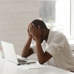Stressed black businessman in panic after business failure sitting at office work desk in front of laptop